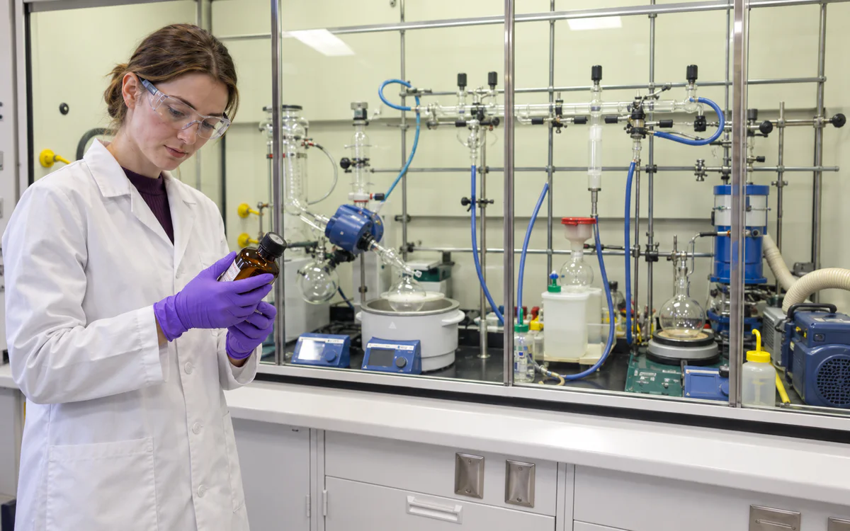 Chemist reviewing a reagent bottle beside laboratory synthesis apparatus.